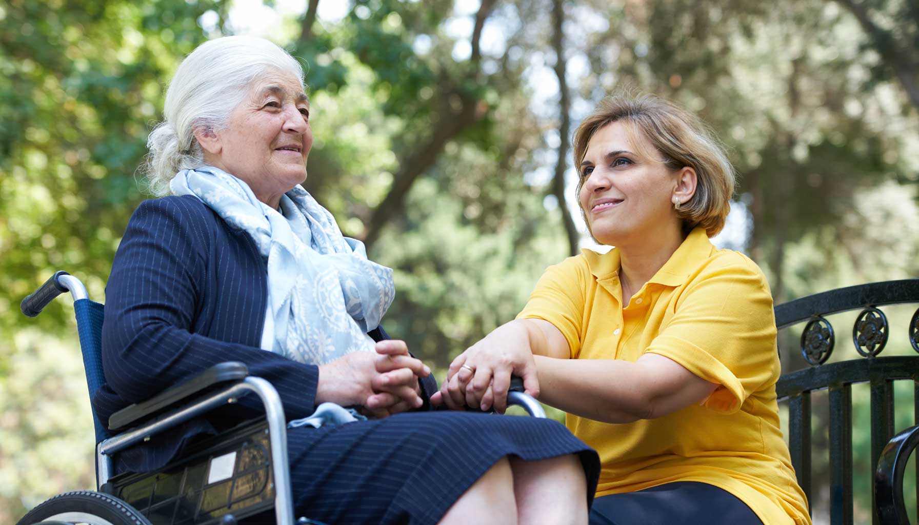 An adult granddaughter sits on a park bench, enjoying a cool, sunny day with her elderly grandmother in a wheelchair. The grandmother, dressed in a coordinating scarf and dress, smiles as she looks out over the scenery.