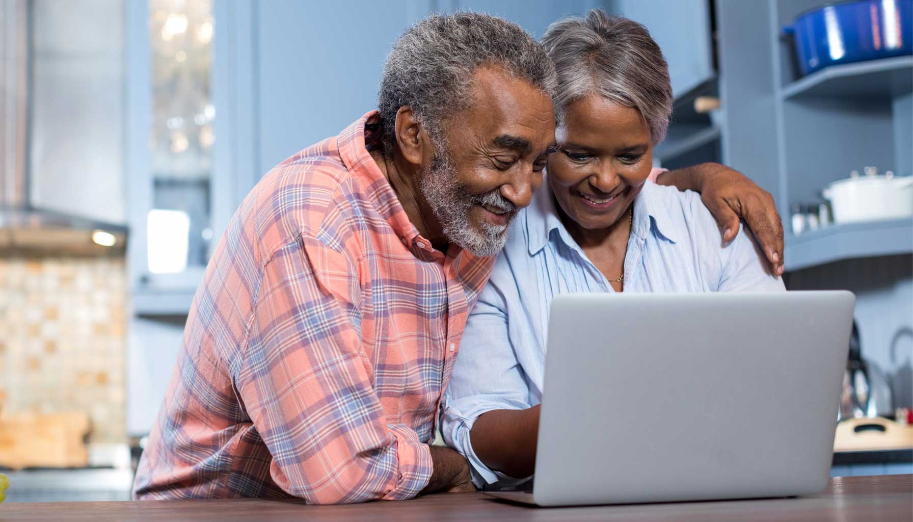 A senior couple researches home care on their laptop. They sit at a wooden table with a modern kitchen visible in the background, complete with a stainless steel stove and restaurant-style fume hood and a professionally installed tile backsplash.